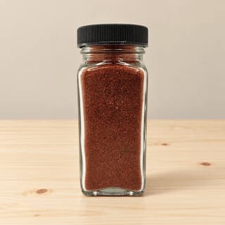Single small clear glass jar of deep red ground sweet paprika with two whole dried chillies resting beside it on pale pine