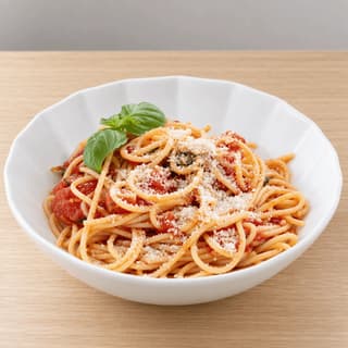 Single white fluted bowl of spaghetti coated in thick tomato sauce with whole basil leaves and grated cheese on blonde oak