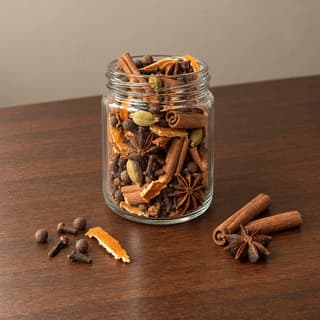 Open plain glass jar of whole mulling spices with cinnamon sticks, star anise, cloves, allspice and dried orange peel on dark walnut