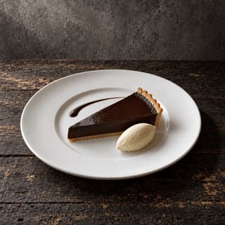 Single wedge of dark chocolate tart with a cream quenelle and a sauce swoosh on a wide bare white rimmed plate