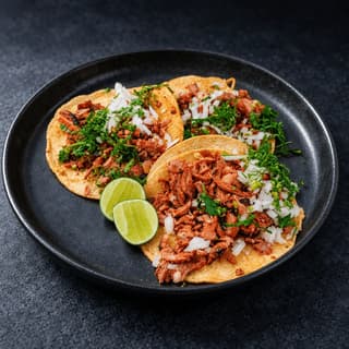 Plate of three tacos al pastor with charred red marinated pork, diced white onion, chopped cilantro and lime halves on a dark plate