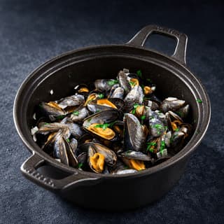 Black cast-iron pot of mussels in white wine and garlic broth with chopped parsley on a dark slate surface