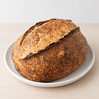 Long scored sourdough batard laid on the diagonal across pale ash wood with a dusting of flour and one wheat stalk