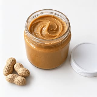 Open unlabelled glass jar of creamy peanut butter with a swirled surface, its plain lid beside it, on a white background with a soft shadow