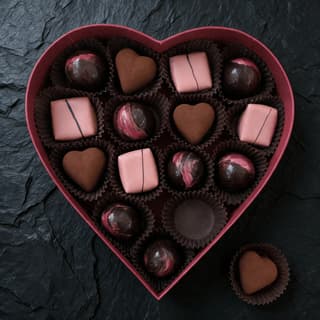 Open heart shaped chocolate gift box photographed from directly above on dark slate, twelve glossy hand finished bonbons seated in fluted brown cups with one cocoa dusted heart resting beside the box