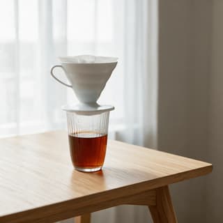 Ceramic pour over cone sitting on a glass carafe with the wet coffee bed domed and blooming and a dark stream falling into the carafe below