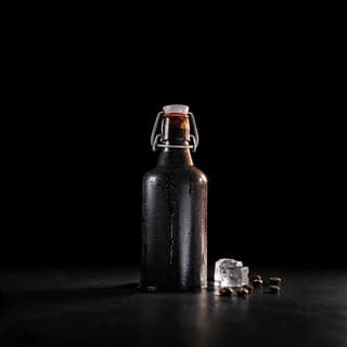 Single unlabelled glass bottle of black cold brew coffee beaded with condensation standing on a jet black surface against a black void