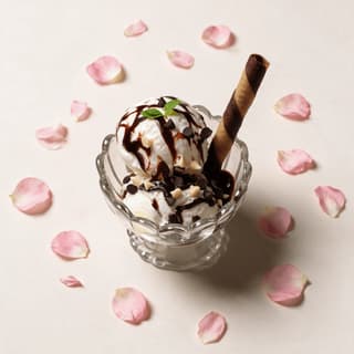 Kem sundae sốt sô cô la nóng trong ly thủy tinh miệng lượn sóng với các viên kem vani, sốt sô cô la đen, vụn sô cô la và một thanh bánh quế cuộn kẻ sọc trên bề mặt màu kem dịu