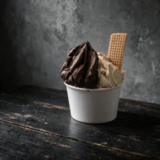 Ly giấy đựng hai vị gelato đặc mịn kèm một chiếc bánh quế, đặt trên quầy gỗ sẫm màu