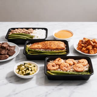 Seasoned salmon fillet over brown rice with asparagus in an open black meal prep container on white marble, with shrimp and ground turkey trays and component bowls around it
