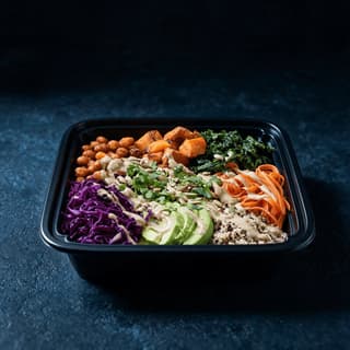 Plant based meal of roasted chickpeas, sweet potato, kale, quinoa, red cabbage, carrot ribbons and avocado with tahini dressing portioned into an open black meal prep container on dark slate