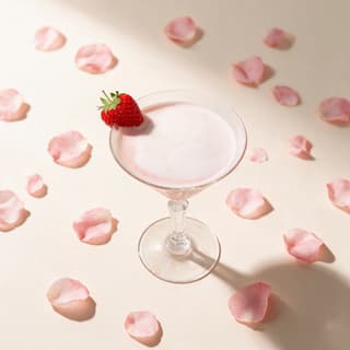Pale blush foamed signature cocktail in a stemmed glass with a fresh strawberry on the rim, on a blush surface scattered with rose petals