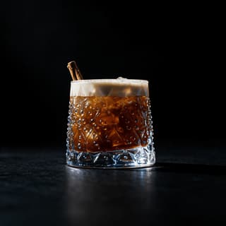 Dark coffee cocktail in a cut crystal tumbler with a pale foam head and a cinnamon stick, on black slate