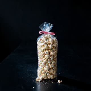 Tall clear bag of birthday-cake popcorn with white drizzle and rainbow sprinkles on a soft pink surface