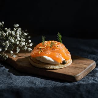 Single canape of smoked salmon and creme fraiche on a small blini topped with dill and lemon zest on a wooden serving board