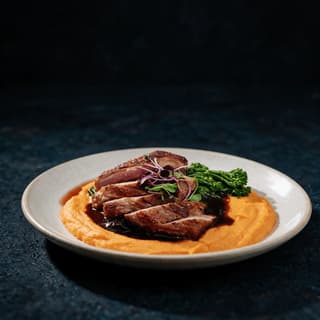 Sliced seared duck breast fanned over a dark cherry reduction and sweet potato purée on a pale rimmed plate under dramatic side light