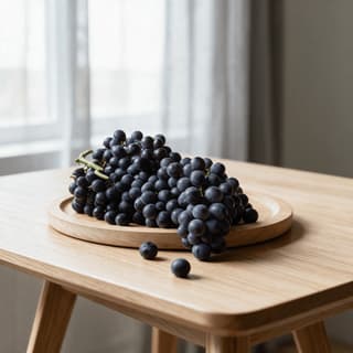 Cluster of deep purple wine grapes with dusty bloom on a shallow round wooden board on a pale oak table in soft window light