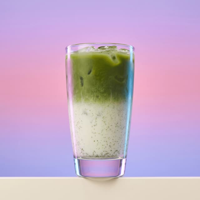 Matcha latte gelado em camadas num copo transparente sobre um degradé lavanda — fotografia de bebidas