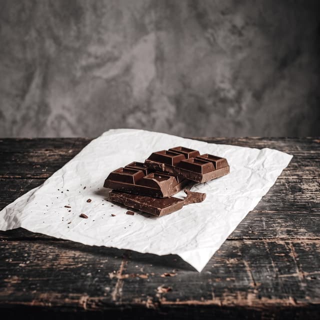 Snapped dark chocolate bar on parchment over weathered dark wood — bean-to-bar chocolate photography