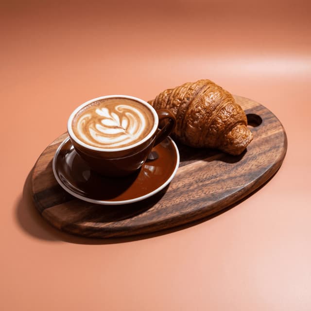 Cappuccino and butter croissant on a walnut board over peach — café pairing photography