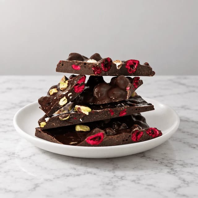 Leaning stack of dark chocolate bark shards with pistachios and dried cherries on a small white plate over white marble