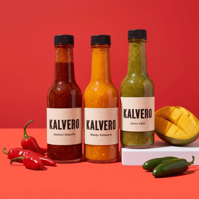 Three hot sauce bottles with fresh chilies and mango on red — product launch poster photography