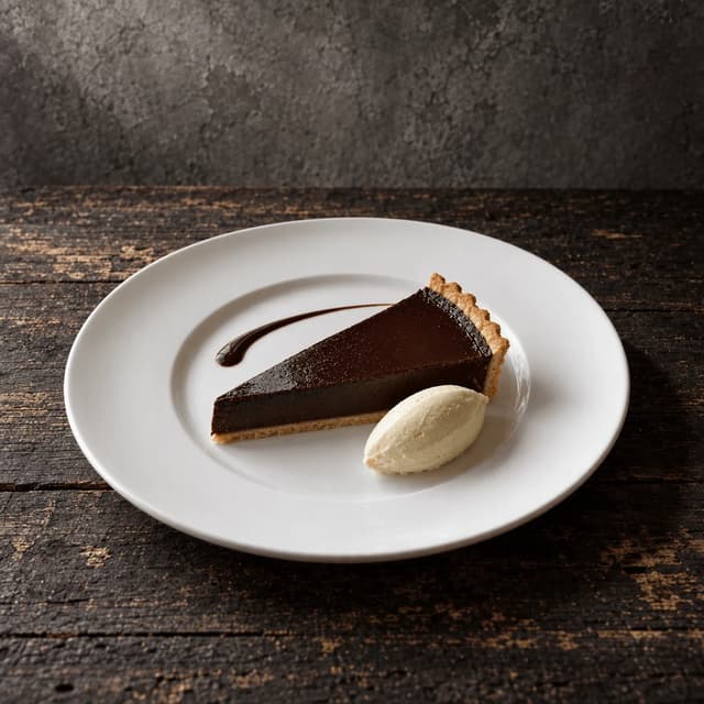 Single wedge of dark chocolate tart with a cream quenelle and a sauce swoosh on a wide bare white rimmed plate