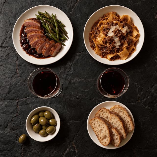 Overhead of a dinner table for two on dark slate with seared duck breast, pappardelle ragu, two glasses of red wine, sliced sourdough and olives