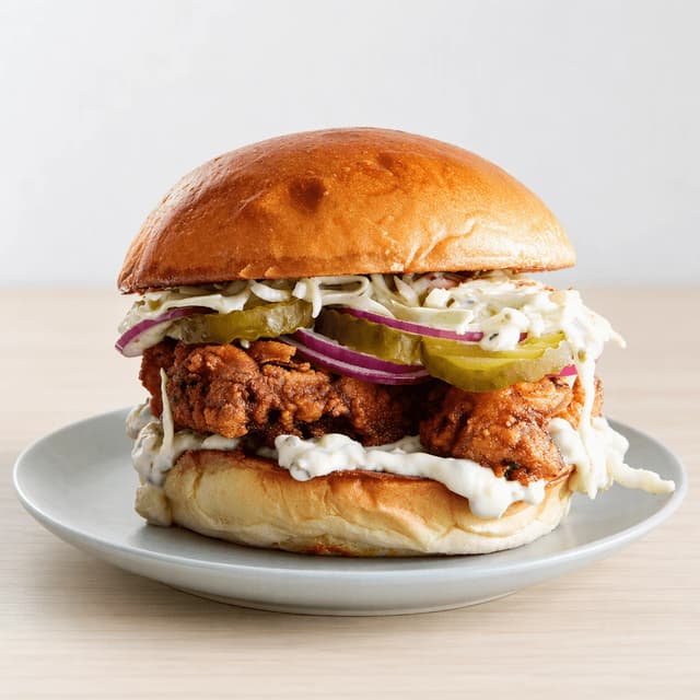 Single buttermilk fried chicken sandwich with pickles, red onion and slaw on a toasted brioche bun on a pale grey plate on pale birch wood