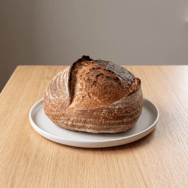 Crusty sourdough boule with a scored ear on blonde oak — small-batch bakery product photography