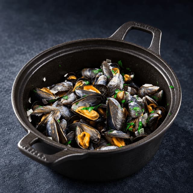 Black cast-iron pot of mussels in white wine and garlic broth with chopped parsley on a dark slate surface