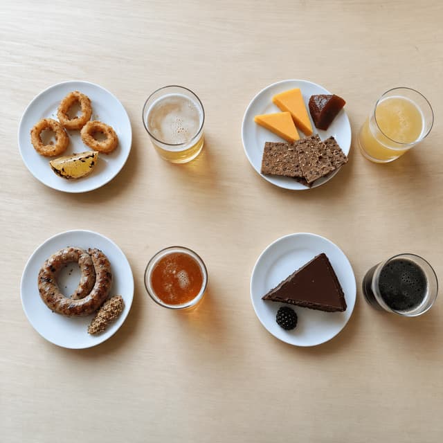 Overhead view of four small pairing plates each set beside a small taster glass of a different beer on pale wood