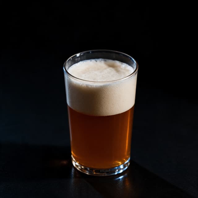 Tall glass of amber craft ale with a thick white foam head and condensation on the glass, lit against a black background