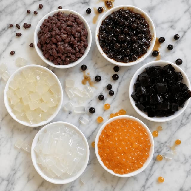Overhead spread of small bowls holding tapioca pearls, grass jelly cubes, popping boba and aloe pieces