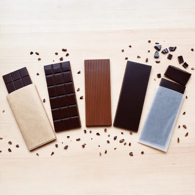 Five single origin chocolate bars fanned out from above with unprinted wrappers and scattered cocoa nibs on pale blond wood