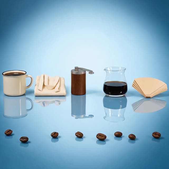 Row of coffee merchandise and brewing equipment including a plain enamel mug canvas tote hand grinder and glass carafe on a glossy blue surface