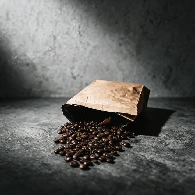 Blank kraft coffee bag tipped on its side on dark grey concrete with roasted beans spilling from the open top in a loose fan