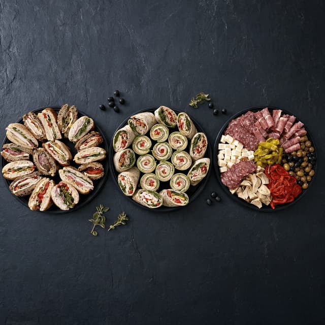 Three separate catering platters of sandwiches wraps and antipasto arranged apart on dark slate photographed from directly above