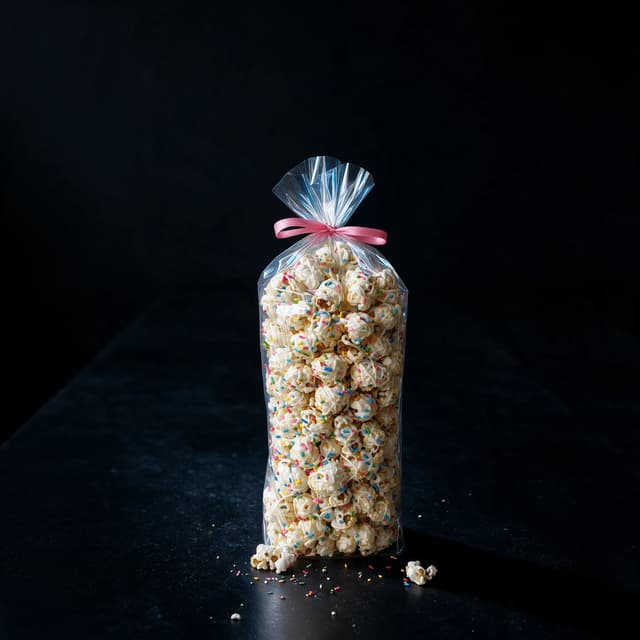 Tall clear bag of birthday-cake popcorn with white drizzle and rainbow sprinkles on a soft pink surface