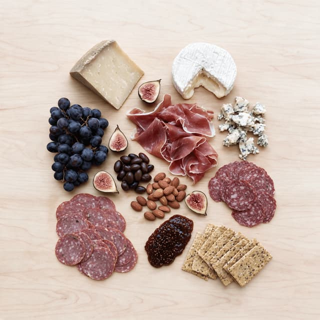 Large grazing board loaded with cheeses, cured meats, figs, grapes, olives and crackers shot from directly overhead on pale wood