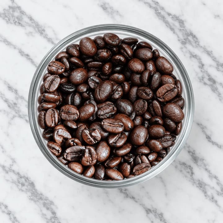 Overhead close-up looking straight down into an unlabelled clear glass container packed with oily dark roasted coffee beans on white marble