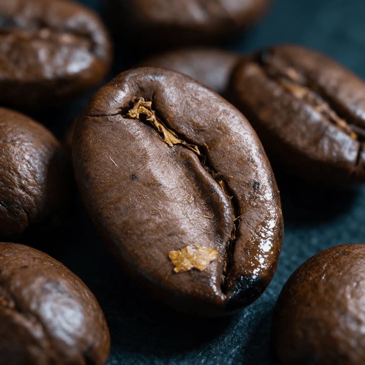 Extreme macro close up of three roasted coffee beans showing the pale centre crack a chaff fleck and a low oil sheen on dark slate
